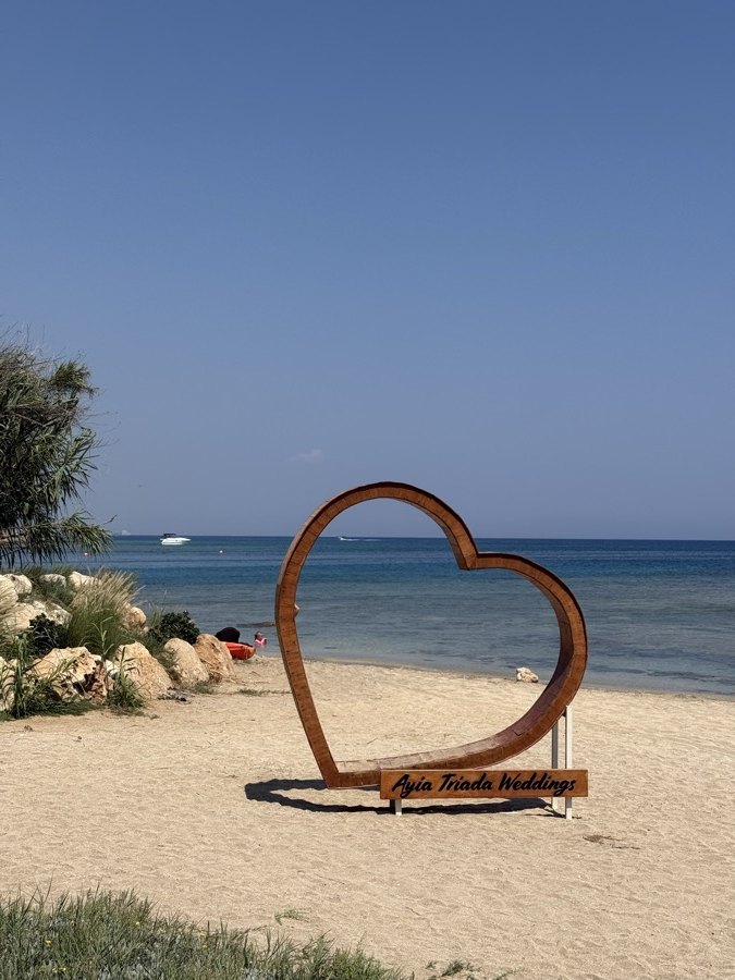 A rusted steel heart sculpture standing on the sand at Kapparis, the sea behind it